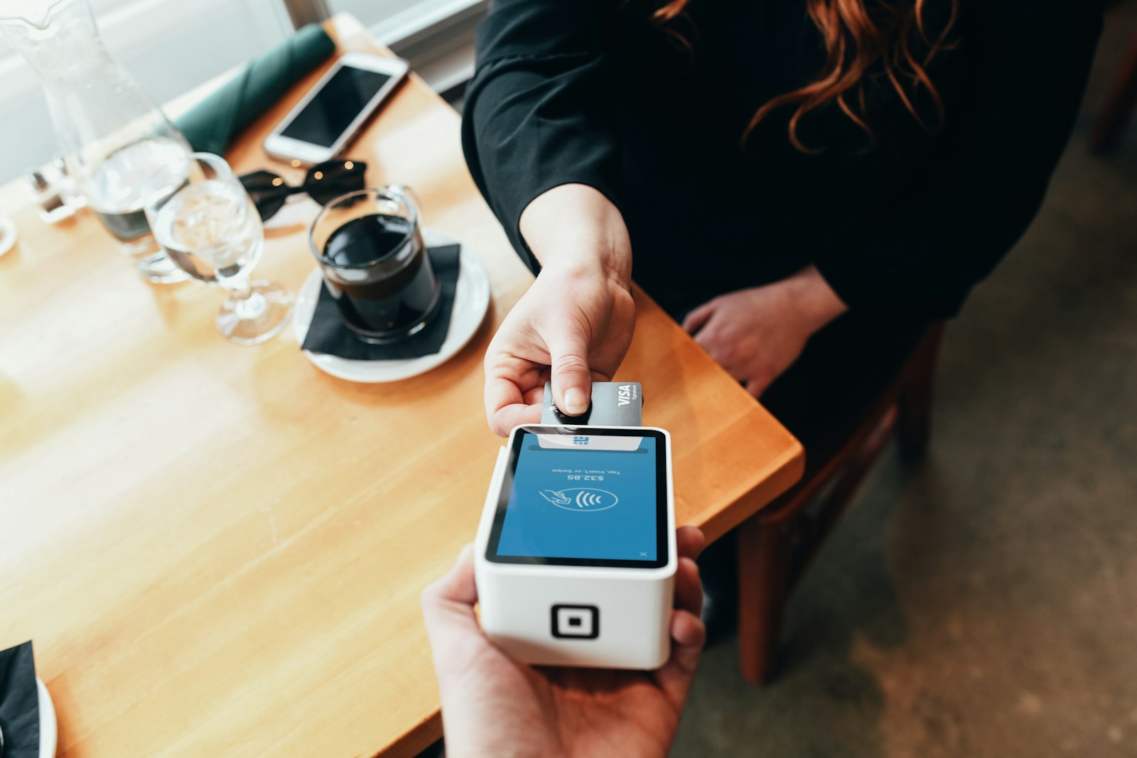Pessoa aproximando um cartão de uma maquininha de pagamento sobre uma mesa. Foto: Blake Wisz / Unsplash
