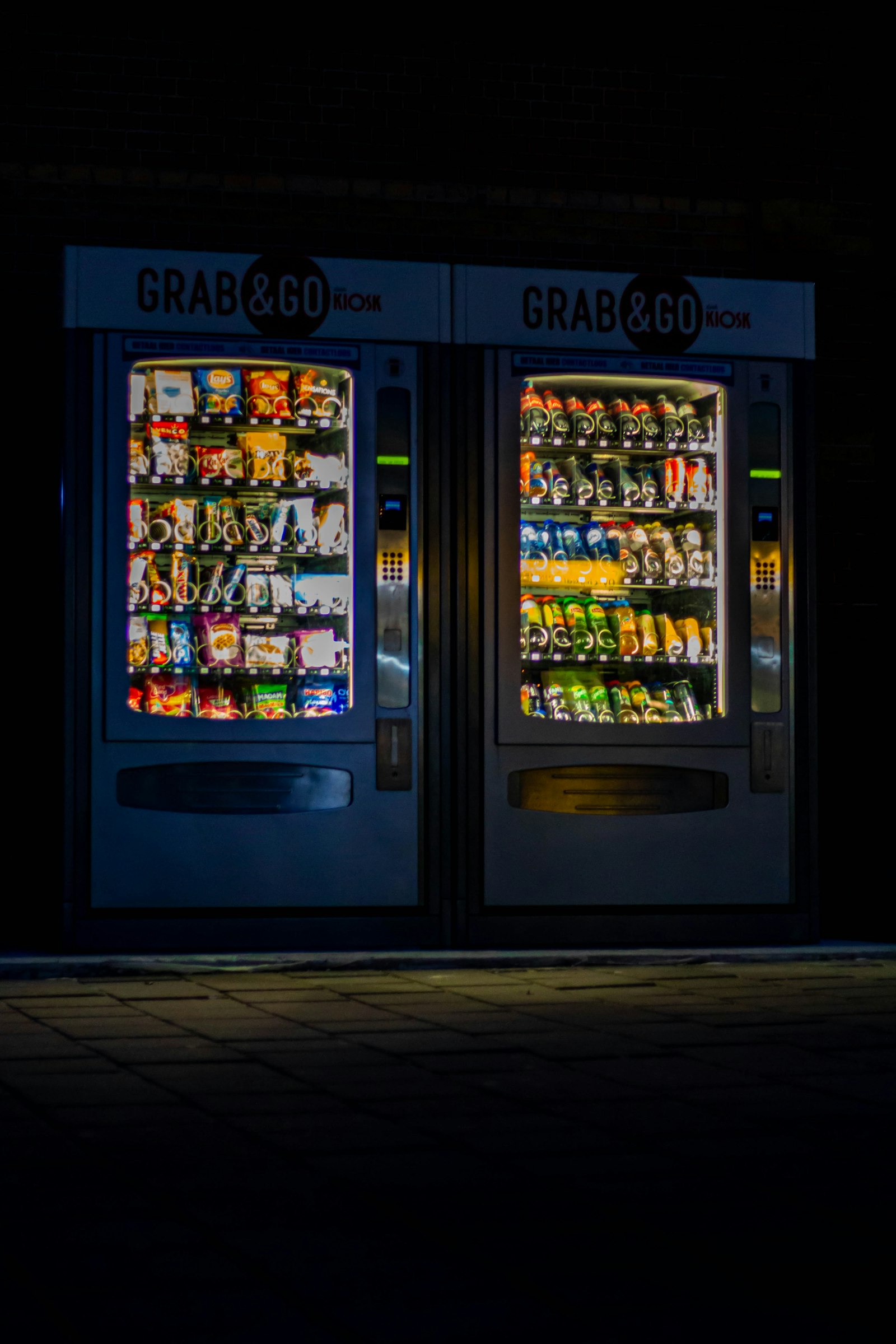 Duas vending machines iluminadas à noite, com produtos visíveis atrás do vidro. Foto: Denise Jans / Unsplash